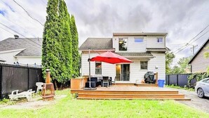 Une maison à deux étages avec un porche à l’avant, un parasol rouge et un grand arbre dans le jardin.