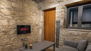 A stone-walled room with a flat-screen TV displaying 'NETFLIX,' a wooden door, a tall floor lamp, a gray sofa, and a small table with a candle.