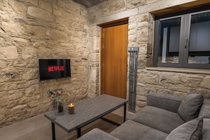 A stone-walled room with a flat-screen TV displaying 'NETFLIX,' a wooden door, a tall floor lamp, a gray sofa, and a small table with a candle.