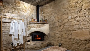 A stone-walled room with a fireplace, a drying rack with white robes, and a bed with a brown blanket.