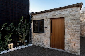 A stone building with a wooden door and window, surrounded by gravel and small plants.