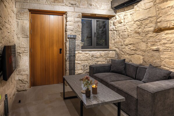 Una habitación con paredes de piedra, una puerta de madera, un sofá gris, una mesa de mármol con una vela y flores, y una ventana con persianas.