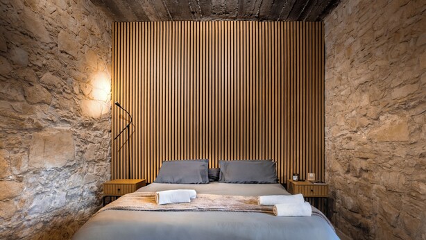 A bedroom with a stone wall, a wooden slat headboard, a bed with grey bedding, two pillows, a bedside table with a lamp, and a towel on the bed.