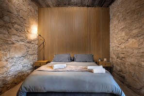Un dormitorio con pared de piedra, cabecera de listones de madera, cama con ropa de cama gris, dos almohadas, mesita de noche con lámpara y una toalla sobre la cama.
