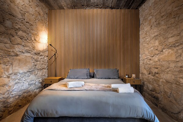 A bedroom with a stone wall, a wooden slat headboard, a bed with grey bedding, two pillows, a bedside table with a lamp, and a towel on the bed.