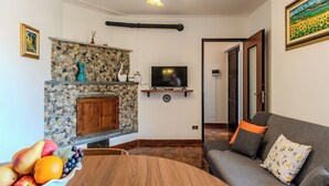 A living room with a stone fireplace, a television, a dining table with fruit, and a sofa with floral cushions. - Cosy apartment for 5 guests with WIFI, patio, pets allowed and panoramic view (Peglio)