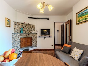 A living room with a stone fireplace, a television, a dining table with fruit, and a sofa with floral cushions.