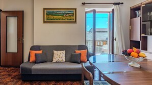 A living room with a sofa, a dining table, a fruit bowl, a painting, and a view of the outside. - Cosy apartment for 5 guests with WIFI, patio, pets allowed and panoramic view (Peglio)