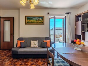 A living room with a sofa, a dining table, a fruit bowl, a painting, and a view of the outside.