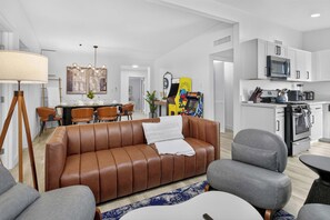 A modern living room with a brown leather sofa, a dining area with a table set for four, and a kitchen with white cabinets and appliances. - 8BR Power Stay | Space, Comfort & Modern Living! (North Las Vegas)