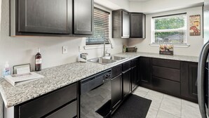 Une cuisine moderne avec des meubles de couleur sombre, des plans de travail en granit, un lave-vaisselle encastré et une fenêtre équipée de stores.