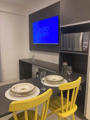 A dining area with a TV displaying 'MODO OREO,' a table with yellow chairs, and a bookshelf with a microwave. - Time Luxury Stay Rooftop premium with sauna and rooftop pool in Maceió (Maceió)