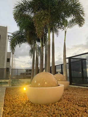 A modern outdoor area with palm trees, a water feature, and a building in the background. - Time Luxury Stay Rooftop premium with sauna and rooftop pool in Maceió (Maceió)