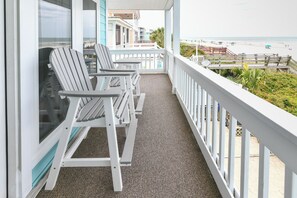 Uma varanda com duas cadeiras Adirondack, guarda-corpo branco e vista para a praia.