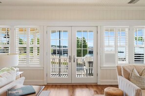 A room with white paneled walls, a wooden floor, a white sofa, a wicker ottoman, and a view of a pool and greenery through white-framed glass doors. - Harbor Cottage: 4  BR, 3.5  BA Cottage in Harbor Springs , Sleeps 12 (Harbor Springs)