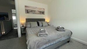 A neatly made bed with a grey checkered bedspread, two pillows, and two folded towels. There is a painting on the wall above the bed and a small bedside table with a lamp. - Tranquil Home Retreat (Berinsfield)