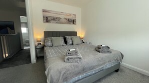 A neatly made bed with a grey checkered bedspread, two pillows, and two folded towels. There is a painting on the wall above the bed and a small bedside table with a lamp. - Tranquil Home Retreat (Berinsfield)