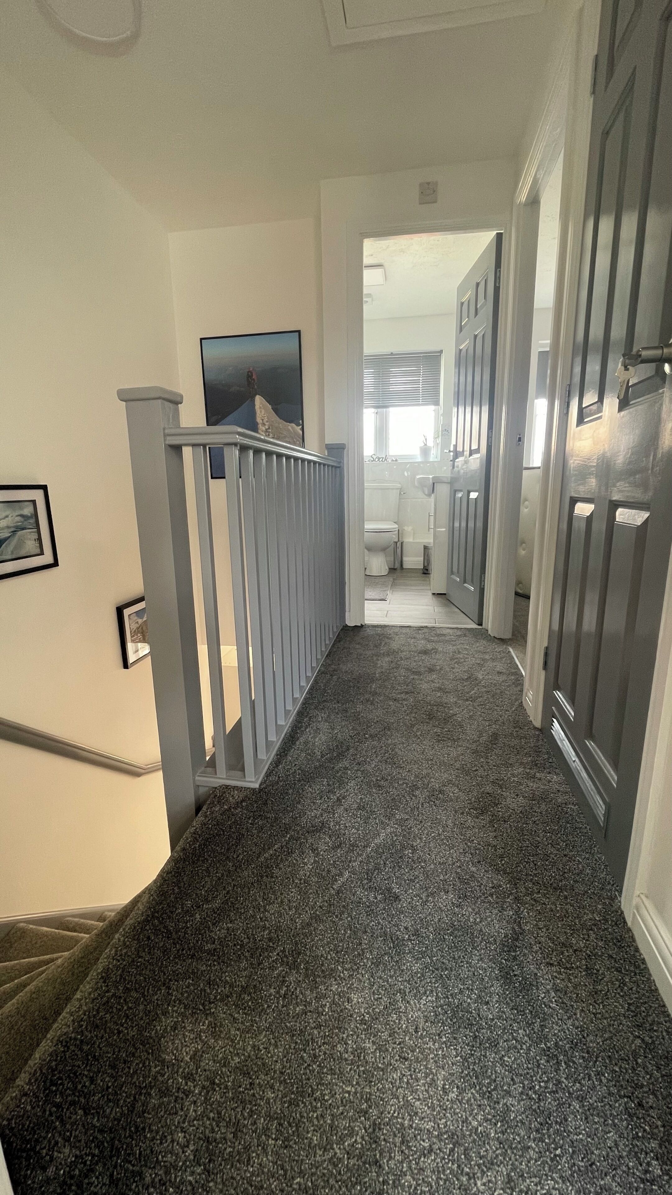 A hallway with a carpeted floor, a staircase with a railing, and a door leading to a bathroom.