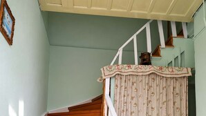 Uma escada de madeira com corrimão branco, uma cortina florida, uma pequena mesa de madeira e um chão de azulejos.