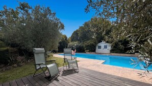 Um deck de madeira com duas espreguiçadeiras voltadas para uma piscina, cercado por árvores e um pequeno galpão.