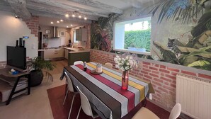 Uma sala de jantar com uma mesa comprida, cadeiras, uma televisão e uma cozinha ao fundo.