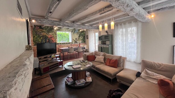 Uma sala de estar com sofá, mesa de centro e televisão. Há um mural na parede e uma janela com cortinas.