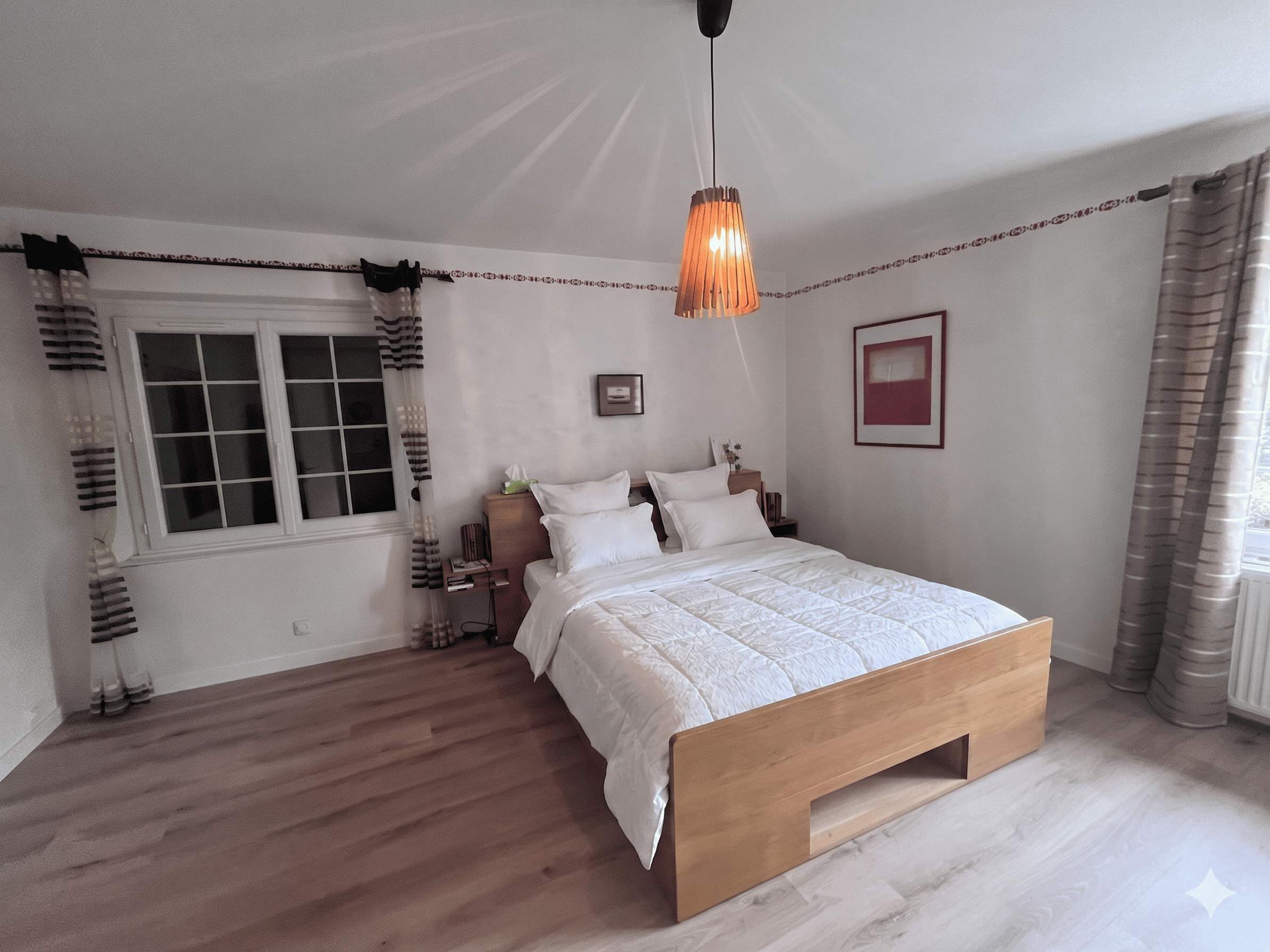 Une chambre à coucher avec un lit en bois, une table de chevet, une commode, un luminaire suspendu, une fenêtre avec des rideaux et un tableau encadré au mur.