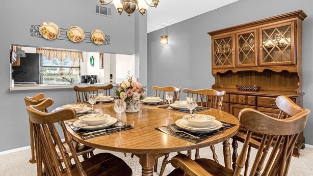 Een eetkamer met een ronde houten tafel, gedekt voor zes personen, omringd door houten stoelen. Een kroonluchter hangt aan het plafond, en aan de rechterkant staat een houten kast met glazen deuren.