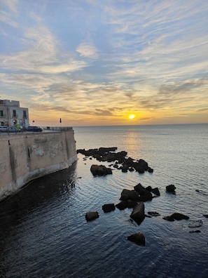 Eine Küstenlandschaft mit Sonnenuntergang, felsigem Ufer und einem Gebäude mit Balkon mit Blick auf das Meer.