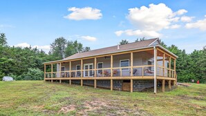 A two-story wooden cabin with a balcony, surrounded by a grassy field and trees. - Dog Friendly, Beautiful Views, Hot tub! (Rileyville)