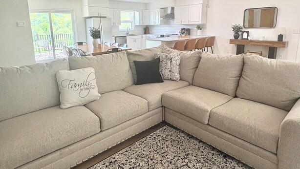Una sala de estar con un sofá seccional color beige, una alfombra estampada y una cocina al fondo.