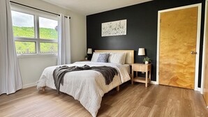 Un dormitorio con piso de madera, una cama con manta blanca y gris, una mesita de noche con lámpara, una ventana con cortinas y una puerta.