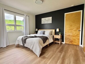 Quarto com piso de madeira, cama com cobertor branco e cinza, mesa de cabeceira com abajur, janela com cortinas e uma porta.