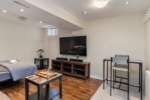 A modern living room with a flat-screen TV, wooden flooring, a bed, a coffee table, and two bar stools. - Walk-out Studio Basement Apartment (Mississauga)