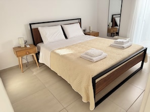 Ein ordentlich bezogenes Bett mit einer beigen Steudecke, zwei Kissen und gefalteten Handtüchern auf einem Bettgestell aus Holz in einem Zimmer mit Fliesenboden.