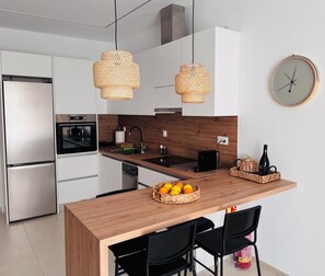 Eine moderne Küche mit weißen Schränken, einem Edelstahlkühlschrank, einer Holzinsel, schwarzen Stühlen und einem Obstkorb.