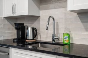 Cuina amb armaris blancs, tauleig negre, cafetera, bullidor, aixeta i ampolla de detergent per plats.
