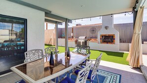 Una terrazza esterna con tavolo in vetro, sedie blu imbottite, una griglia per barbecue e un muro decorativo.