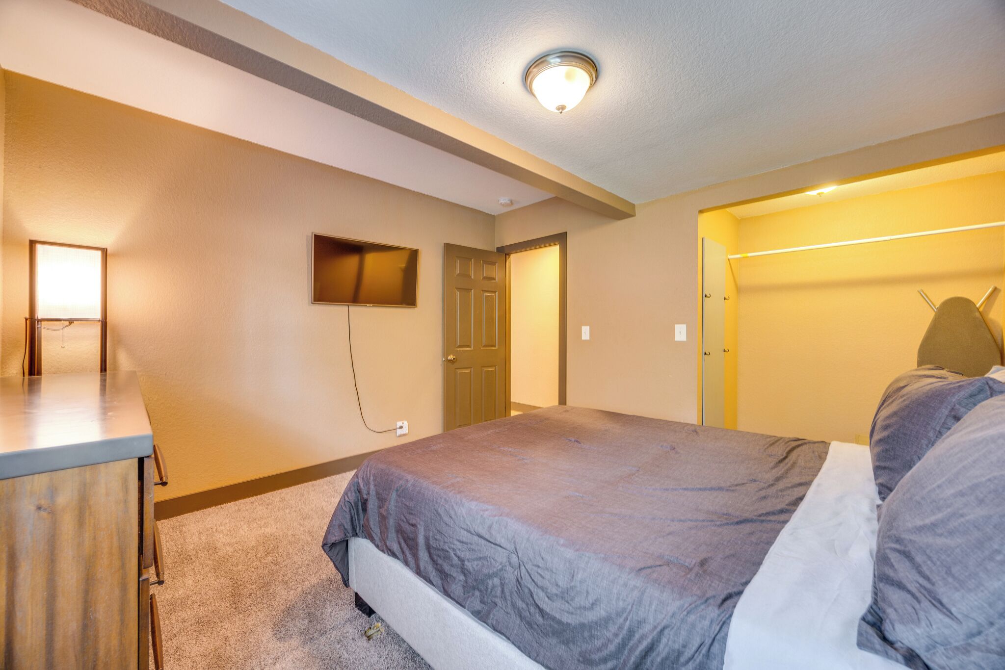 A bedroom with a bed, a desk, a TV mounted on the wall, and a door leading to another room.