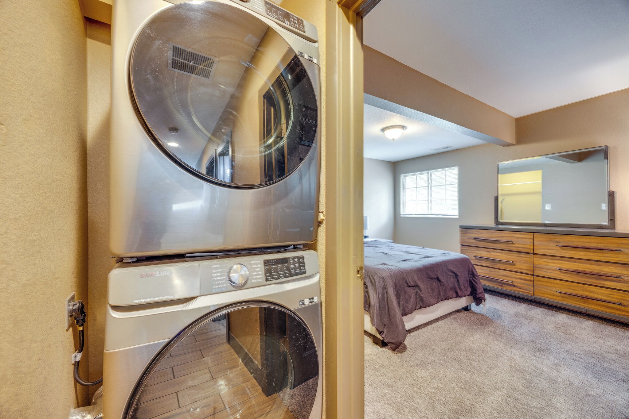 A laundry room with a stacked washer and dryer, a bed with a brown blanket, a dresser, and a large mirror.
