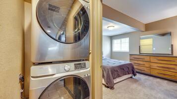 A laundry room with a stacked washer and dryer, a bed with a brown blanket, a dresser, and a large mirror.