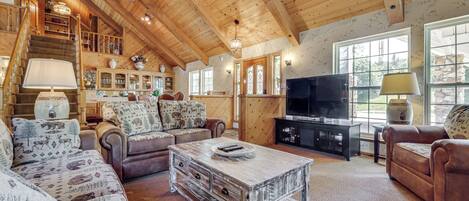 A spacious living room with a wooden ceiling, a fireplace, a television, and a staircase leading to an upper level.