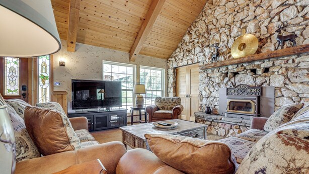 A cozy living room with a stone wall, wooden ceiling, leather sofas, a fireplace, and a television. - 25-acre Hideaway in Oakhurst: Pet Friendly! (Oakhurst)