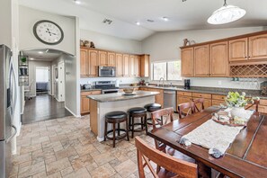 A kitchen with wooden cabinets, a central island with stools, a dining table with chairs, and a clock on the wall. - Spacious Backyard! Family Haven in San Tan Valley (San Tan Valley)