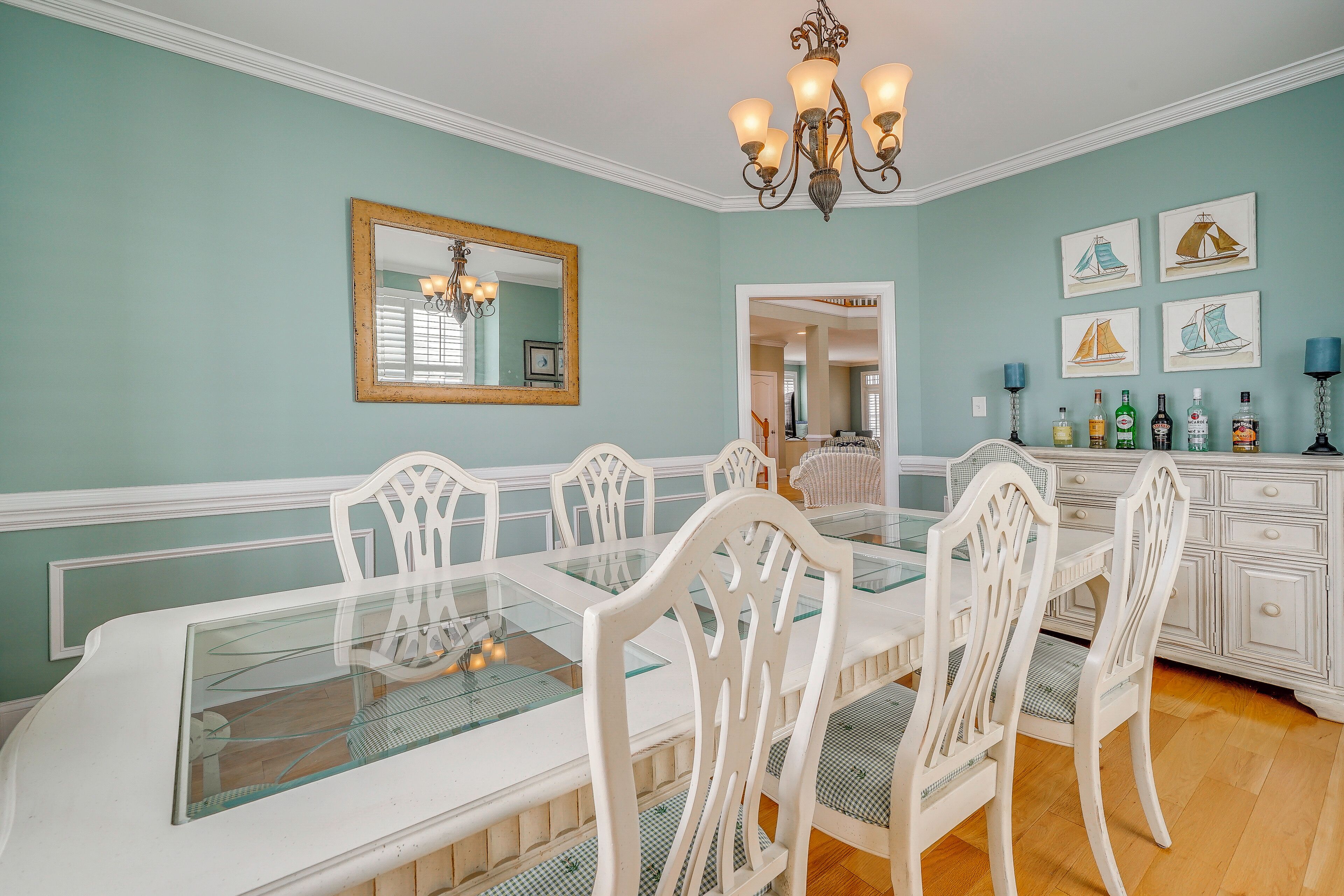 A dining room with a glass-top table, white chairs, a chandelier, a mirror, and wall art.