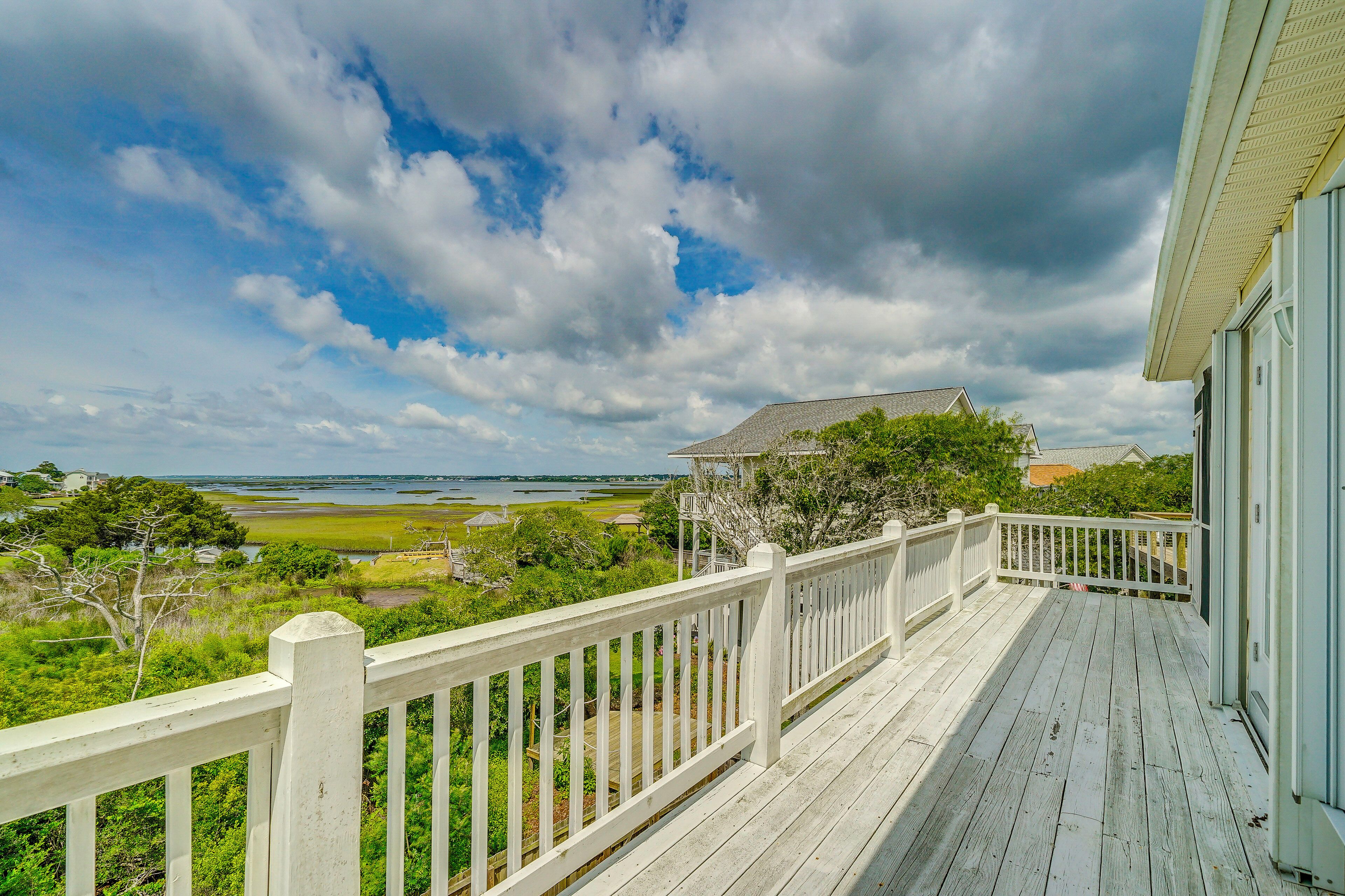A wooden deck with a white railing overlooking a coastal landscape with a body of water and greenery.