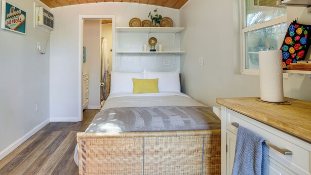 A bedroom with a bed, a wicker chair, a wooden ceiling, a window, and a bathroom visible in the background. - Tiny House Experience in Las Vegas w/ Free Parking (Las Vegas)