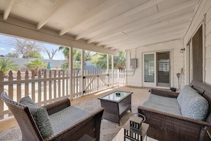 A covered patio area with wicker furniture, a glass table, and a view of the backyard through a white fence.