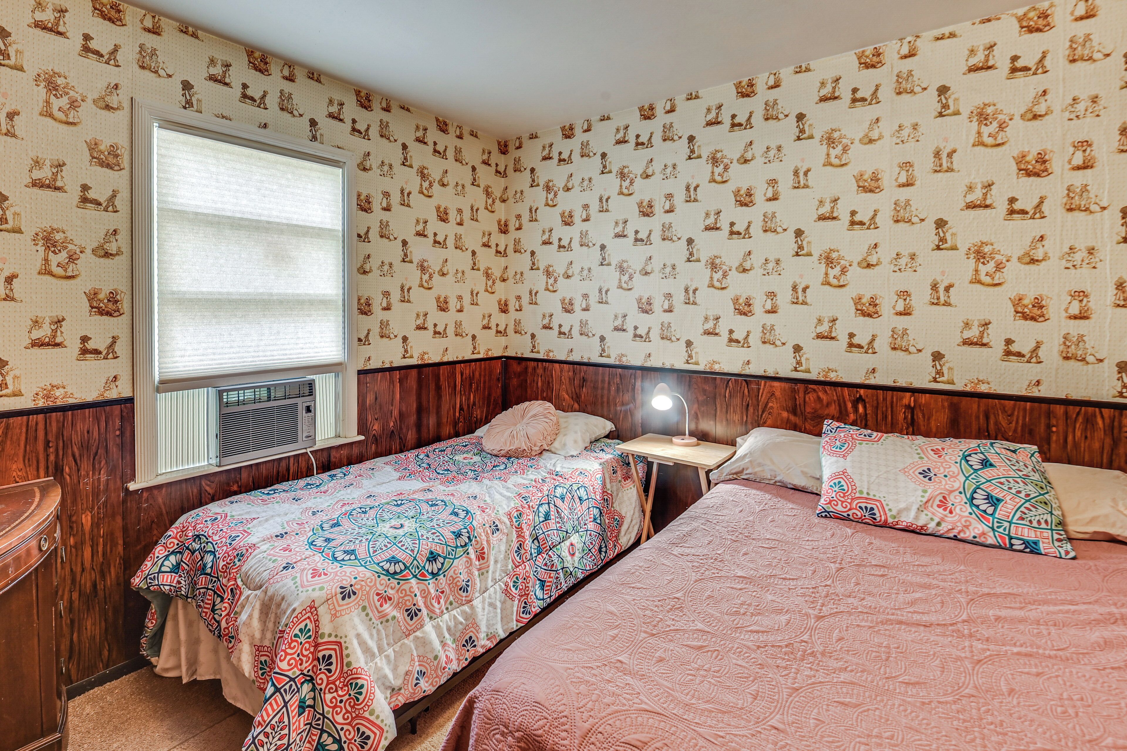 Ein Schlafzimmer mit zwei Betten, einem Fenster mit Jalousien, einem Nachttisch mit Lampe und Wandtapete mit Muster.