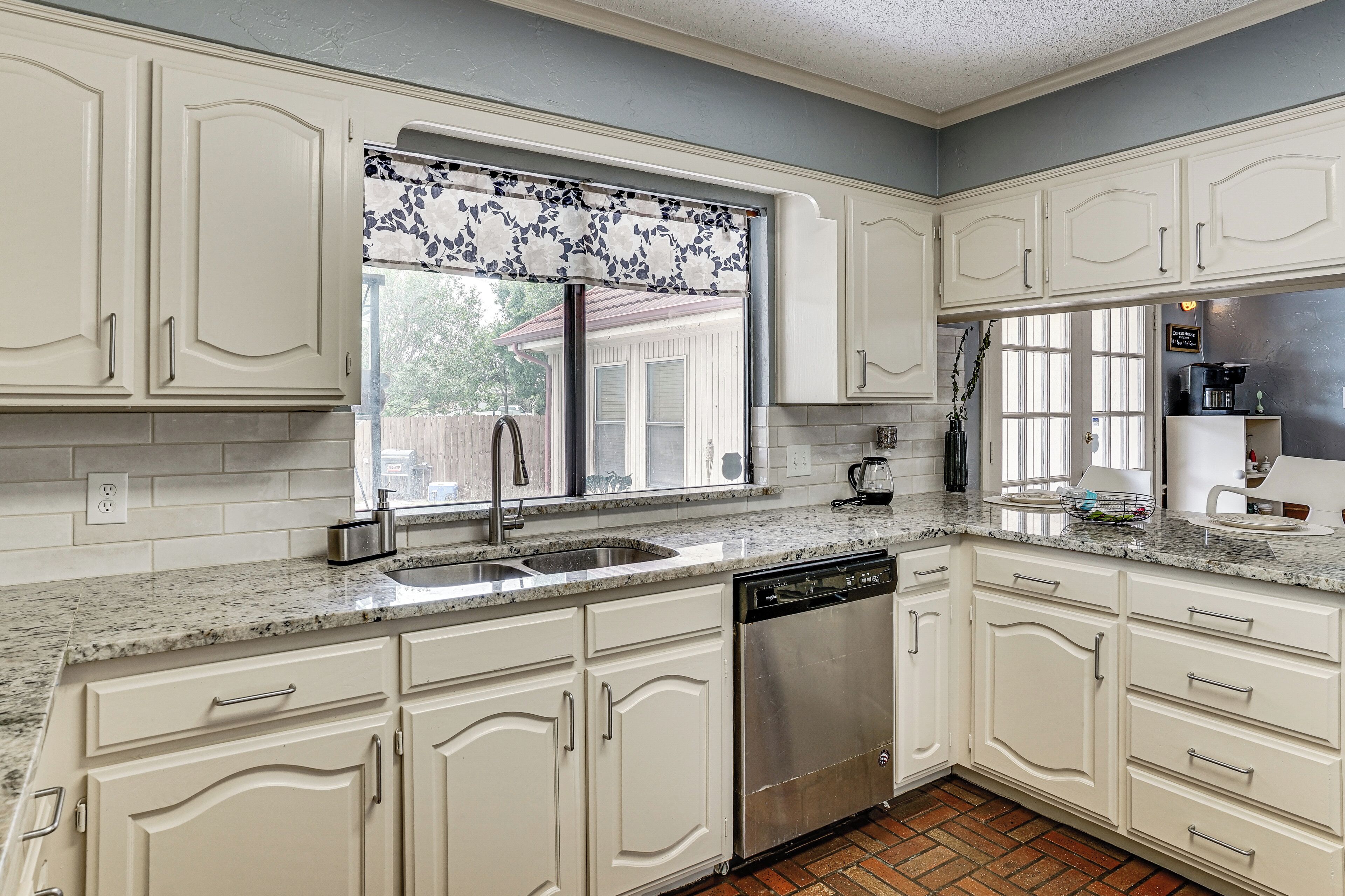 Cozinha com armários brancos, lava-louças de aço inoxidável e janela com cortina decorada.